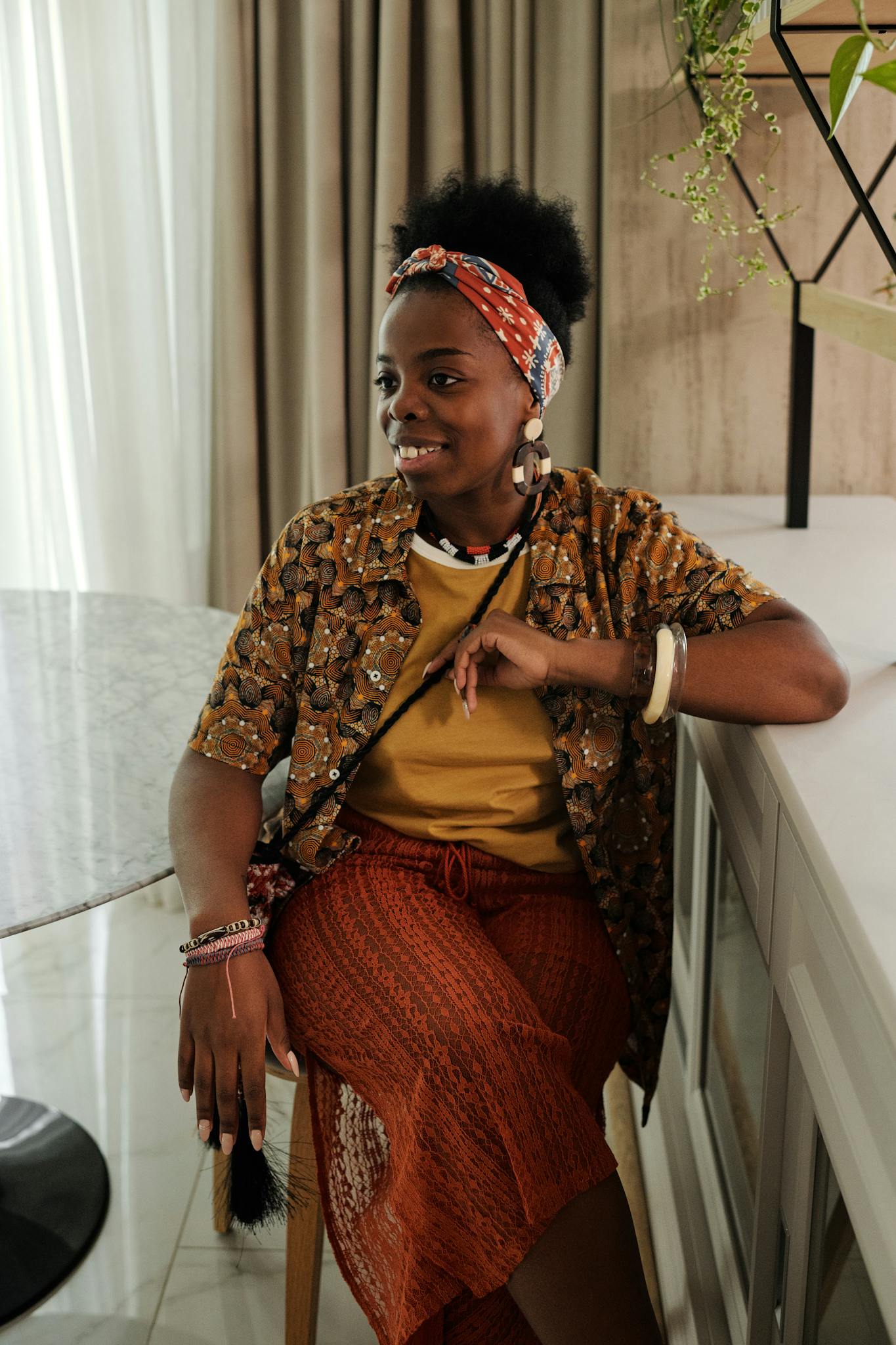 Cheerful woman sitting indoors, wearing fashionable outfit with a headband and jewelry.